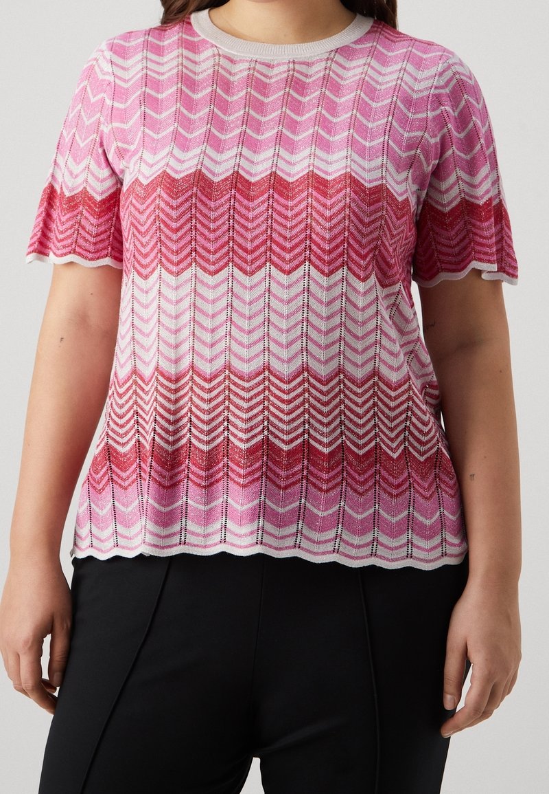 Svetr s krátkým rukávem pletený v růžovo-červeném chevron vzoru, se strukturovanými vlákny a vlnitým lemem, doplněný bílým obrysem.