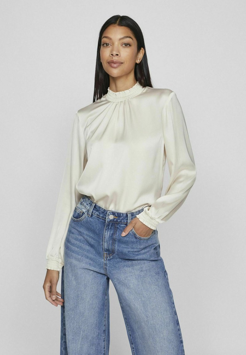 Blouse en satin crème avec un col froncé haut et des manches longues. Associée à un jeans large bleu clair avec patte de boutonnage et poches latérales.