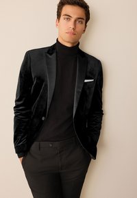 Black velvet blazer with satin lapels, worn over a black turtleneck. Slim-fit black trousers. Notable pocket square detail.