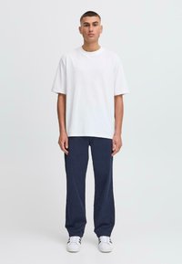 Hvid bomulds t-shirt med korte ærmer, parret med navy blå afslappede bukser. Modellen står oprejst med hvide sneakers. Minimalistisk stil.