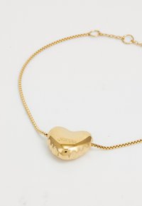 Gull hjerteformet armbånd med teksturert overflate, utstyrt med en glatt kjede. "GUESS"-logoen er subtilt gravert på hjertet.