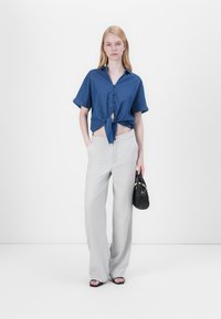 Denim overhemd met korte mouwen, knoopsluiting en geknoopt voorpand, gecombineerd met lichtgrijze wijde broek. Model houdt een kleine zwarte handtas vast.