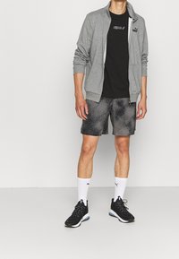 Pánský outfit Puma se šedou zapínací bundou, černým tričkem, vzorovanými šedými šortkami, bílými ponožkami a černými sportovními botami.