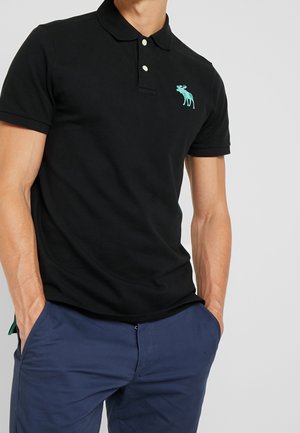 Mężczyzna w czarnej koszulce polo z krótkim rękawem z turkusowym logo łosia i granatowych spodniach, ręce w kieszeniach, na jednolitym tle.