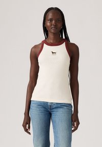 Kremfarget tank topp med burgunder kanter og ribbestruktur. Har et lite brodert dachsdesign på brystet. Kombinert med blå jeans.