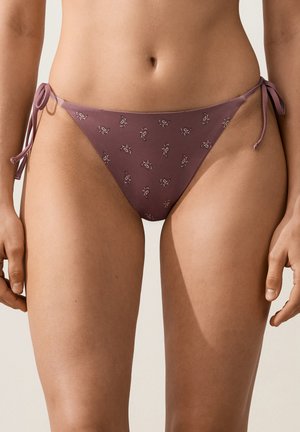 Zbliżenie osoby noszącej majtki bikini w kolorze fioletowym z małymi wzorami paisley i wiązaniami po bokach, ukazujące dolną część brzucha i górną część ud.