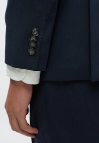 Tengerészkék, texturált zakóujj fekete gombokkal, alatta egy tiszta fehér ingmandzsetta látható.