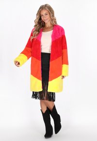 Manteau en fausse fourrure arborant un motif en chevrons dans des couleurs vives : rose, rouge, orange et jaune. Assorti à une jupe noire à franges et des bottes noires.