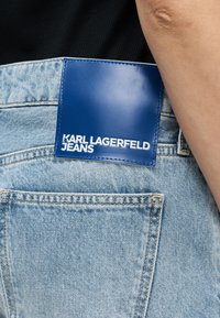 Τσέπη πίσω από ανοιχτό μπλε τζιν με μπλε δερμάτινη ετικέτα που γράφει "KARL LAGERFELD JEANS" σε άτομο που φοράει μαύρο τοπ.