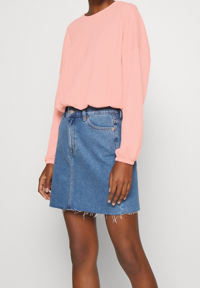 Vaaleanpunainen teksturoitu pitkähihaisten paita, yhdistettynä keskipitkään siniseen farkkuhameeseen, jossa on rikkinäinen helma ja klassinen viiden taskun muotoilu.