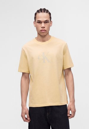 Junger Mann mit geflochtenem Haar, der ein beiges Calvin Klein T-Shirt und dunkle Hosen vor einem schlichten hellen Hintergrund trägt.