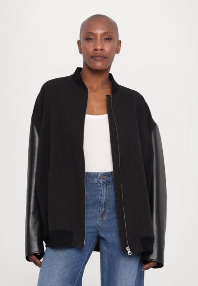 Black bomber jacket with contrasting leather sleeves, zip closure, and a relaxed fit. Worn over a white top and blue jeans.