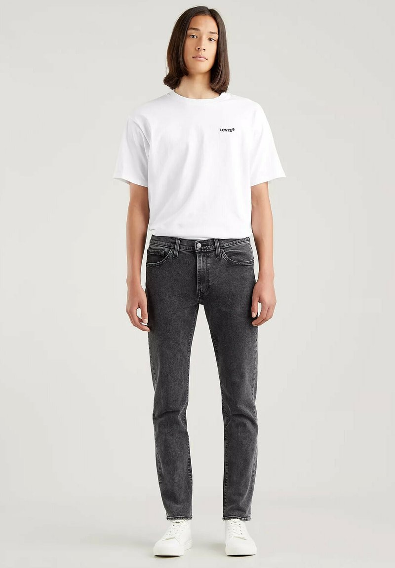 T-shirt bianca a girocollo con logo "Levi's" nero abbinata a jeans scuri aderenti grigi e sneakers bianche, stile complessivo casual e texture liscia.