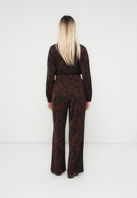 Femme avec des cheveux blonds lisses, portant un haut à fleurs rouge foncé et noir à manches longues et un pantalon large assorti, faisant face à un mur blanc.