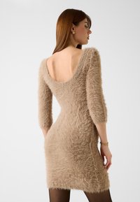 Beige, pluizige jurk met een diepe V-rug en driekwart mouwen; heeft een aansluitende silhouet en zachte textuur. Minimale details.