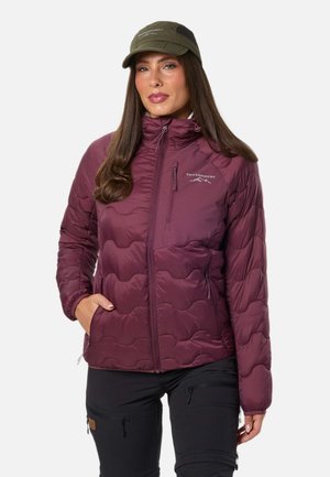 Une femme porte une veste d’extérieur matelassée bordeaux, un pantalon de randonnée noir et une casquette olive, sur un fond blanc uni.