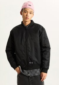 Black bomber jacket with a front zipper, ribbed cuffs and waistband, and a utility pocket on the sleeve. Worn over a checkered shirt.