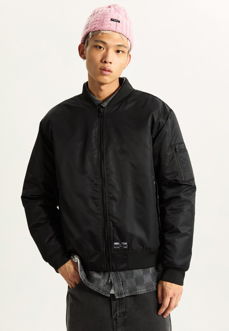 Black bomber jacket with a front zipper, ribbed cuffs and waistband, and a utility pocket on the sleeve. Worn over a checkered shirt.