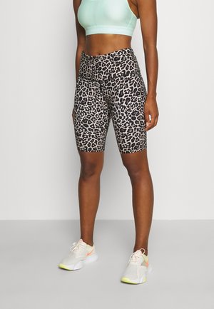 Kvinde iført leopardprint højtaljede biker-shorts, lysegrøn sports-BH og hvide sneakers med neon-accenter, stående mod en ensfarvet baggrund.