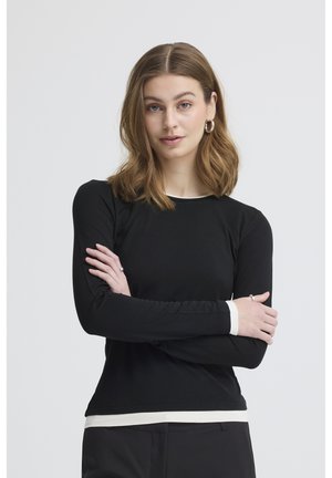 Młoda kobieta o brązowych włosach do ramion, ubrana w czarną koszulę z długim rękawem i białym obszyciem, pozująca z założonymi na siebie ramionami na jednolitym tle.