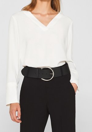 Vrouw die een witte blouse met lange mouwen en V-hals draagt, ingestopt in een zwarte broek met een brede zwarte riem met een grote zilveren ronde gesp.
