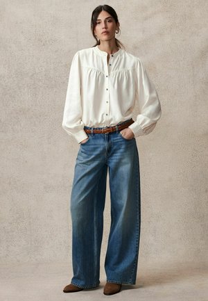 Crème blouse met opgeblazen mouwen en knoopsluiting, gecombineerd met wijde blauwe jeans en een bruine gevlochten riem. Bruine enkellaarsjes maken de look af.