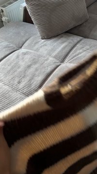 Ugodan, teksturirani sivi sofa s vertikalnim rebrima, opremljen jastučićem u istoj sivoj boji. Djelomično je vidljiv smeđi i kremasti prugasti pokrivač.