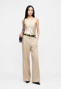 Γυναίκα στέκεται φορώντας μπεζ σατέν τοπ με ρυτιδώσεις, μπεζ παντελόνι με φαρδιά πόδια και μαύρη ζώνη, μαύρα μυτερά παπούτσια και μαύρη τσάντα ώμου.