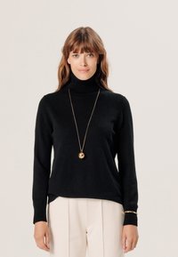 Schwarzer Rollkragenpullover aus weichem Stoff. Besitzt lange Ärmel und einen lockeren Schnitt, kombiniert mit beigefarbenen, taillierten Hosen und einer goldenen Halskette.