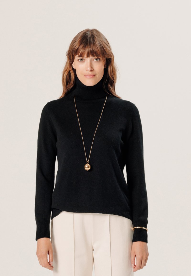 Schwarzer Rollkragenpullover aus weichem Stoff. Besitzt lange Ärmel und einen lockeren Schnitt, kombiniert mit beigefarbenen, taillierten Hosen und einer goldenen Halskette.