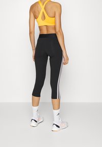 Donna in piedi di spalle che indossa leggings neri a vita alta con strisce bianche, un reggiseno sportivo giallo, calze Adidas bianche e sneakers bianche.