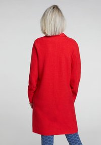 Persona con capelli biondi corti, che indossa un cappotto rosso brillante sopra il ginocchio e pantaloni blu a fantasia, in piedi su uno sfondo chiaro e uniforme.