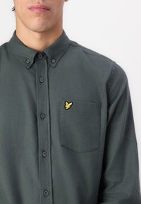 Cămașă butonată de culoare verde închis, dintr-un material moale, cu guler, un buzunar pe piept și un logo cu un vultur brodat în aur pe buzunar.