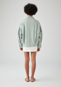 Veste en denim oversize vert clair avec des épaules tombantes, des poignets à bouton et un col, associée à une mini-jupe blanche.