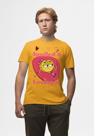 Gul bomulds T-shirt med en tegneseriefigur med en sløjfe, grafisk tekst "Forkælet & Elsker Det" på en pink hjertebaggrund.