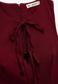 Top senza maniche color bordeaux con scollatura tonda, chiusura a zip frontale e lacci decorativi. Tessuto liscio con una leggera texture.