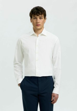 Giovane uomo con capelli ricci e scuri, indossa una camicia bianca con bottoni e pantaloni blu navy, in piedi con una mano in tasca.