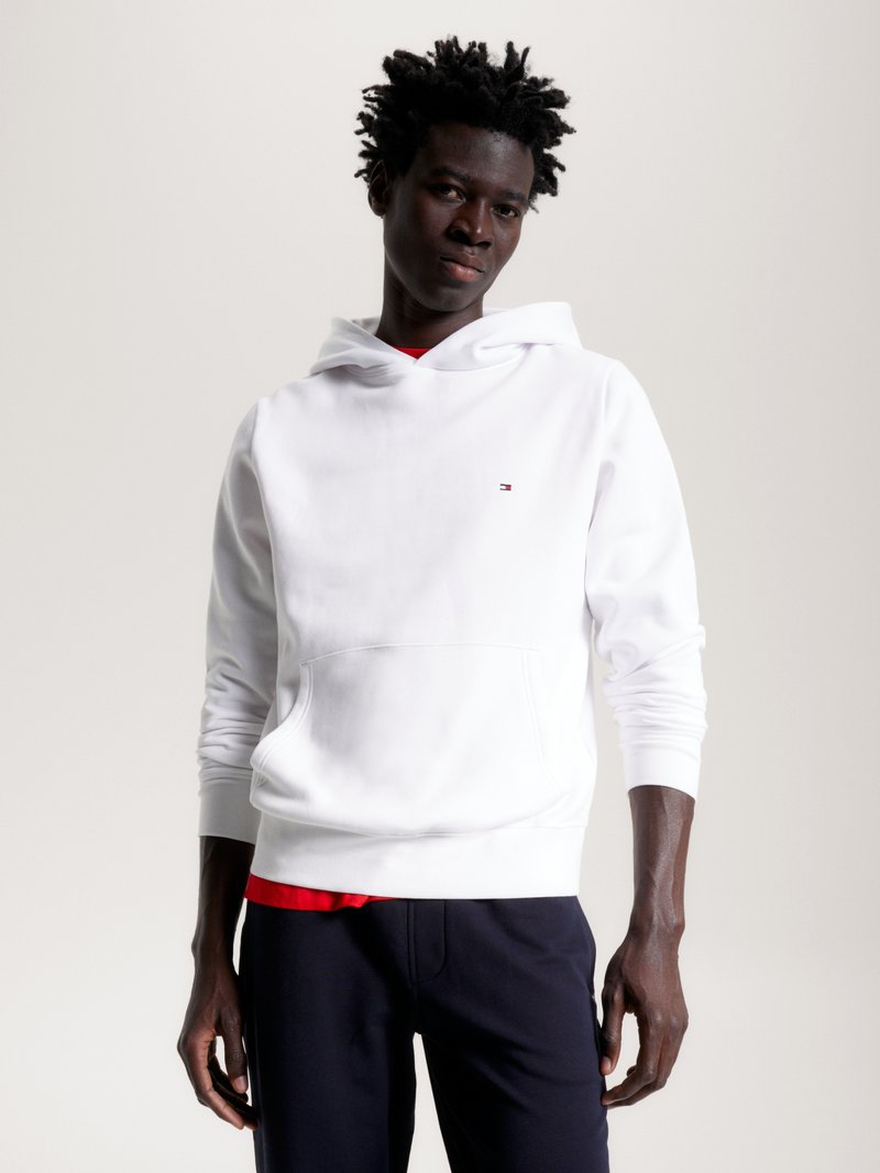Sweatshirt branco com bolso canguru e punhos canelados, sobre uma camisa vermelha, combinado com calças escuras. Apresenta um logo no peito.