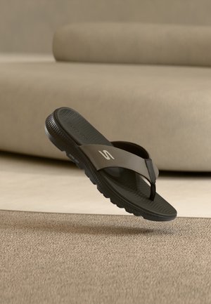 Sandale Skechers noire et grise à lanière entre les orteils avec une semelle texturée flottant au-dessus d'un tapis beige devant un canapé beige.