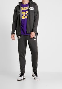 Sudadera gris con cremallera, camiseta morada de los Lakers con el número 23, pantalones jogger grises con el logo de Nike. Conjunto combinado con zapatillas negras y blancas.