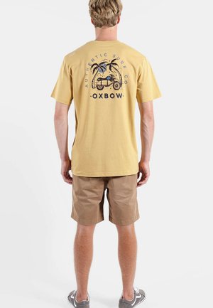 Homme portant un t-shirt jaune avec un motif de moto et de palmier et le texte "Authentic Surf Co Oxbow", assorti à un short marron et des baskets grises.