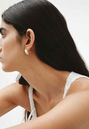 Pendientes en oro y blanco con un diseño ovalado liso y un borde texturizado. Primer plano que resalta el área de la oreja y el cuello. Sencillos y minimalistas.