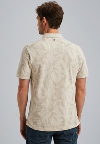Polo beige avec un motif de feuilles subtil, à manches courtes et avec un col. Tissu texturé avec une coupe décontractée, porté avec un jean noir.