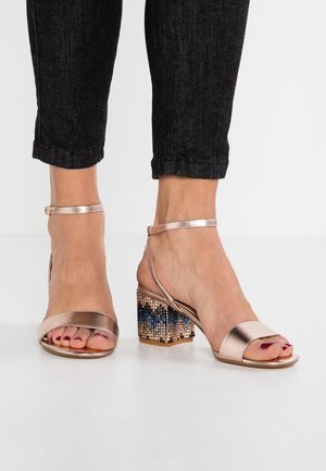 Füße, die metallisch roségoldene Sandalen mit offenen Zehen, Knöchelriemen und Blockabsätzen tragen, die mit mehrfarbigen Strasssteinen verziert sind, kombiniert mit schwarzen verkürzten Hosen.