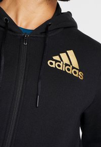 Sudadera con cremallera negra hecha de tejido texturizado, con un logo dorado de Adidas en el lado izquierdo del pecho y cordones en la capucha.