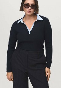 Woman wearing dark sunglasses, navy sweater over light blue collared shirt, and dark pinstriped trousers, standing against plain background.