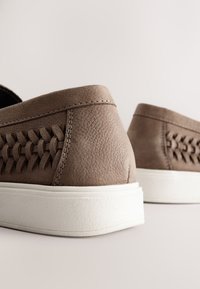 Brown suede sneakers with woven side design, white rubber sole, and textured surface. Stitch detailing along the upper edge.