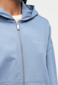 Ανοιχτό μπλε ζακέτα με φερμουάρ φτιαγμένη από μαλακό ύφασμα, διαθέτοντας κουκούλα με κορδόνι και κεντημένο λογότυπο της Calvin Klein στο αριστερό στήθος.