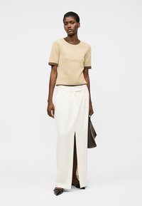 Kvinne iført en beige short-sleeve strikket topp og høytlivs hvit skjørt med frontsplitt, som holder en mørk brun håndveske og svarte spisse sko.