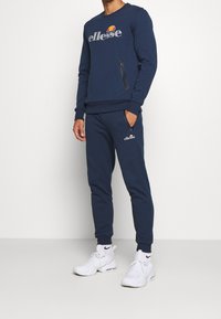 Sweat-shirt bleu marine avec un logo, assorti à un pantalon de jogging. Comprend des poches zippées et des poignets côtelés. Porté avec des baskets blanches.